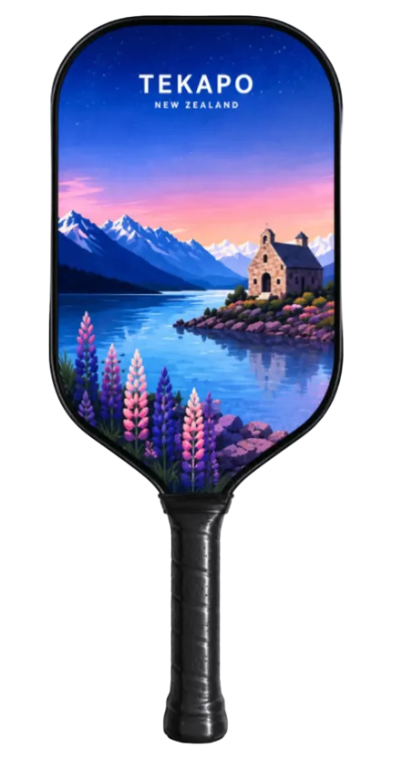 A pickleball paddle featuring a scenic New Zealand print with the text 'TEKAPO NEW ZEALAND' at the top. The paddle face displays a colorful landscape with a church, mountains, a lake, and purple flowers under a blue and pink sky, while the handle is black.
